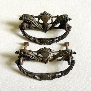 May include: Two antique bronze drawer pulls with ornate, symmetrical designs. Each pull features a curved handle and decorative elements, including a central shield shape and scrolling details. The pulls are mounted with screws.