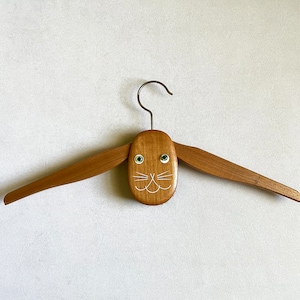 May include: A wooden hanger shaped like a rabbit with a painted face and green eyes. The hanger has a metal hook.