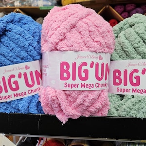 May include: Three balls of super chunky yarn in blue, pink, and green. Each ball has a label that reads "BIG'UN Super Mega Chunky" by James C. Brett. The yarn is displayed on a black shelf.
