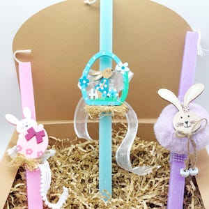 Può includere: Tre candele pasquali in una scatola con carta triturata. Le candele sono decorate con coniglietti, uova e fiori. Le candele sono rosa, blu e viola.