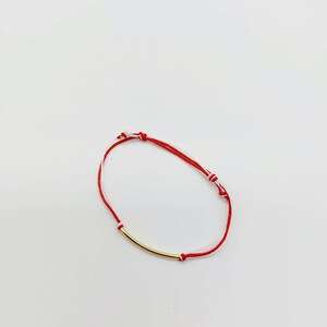 May include: A red and white string bracelet with a gold bar charm.