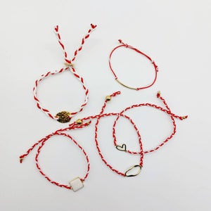 May include: Five red and white string bracelets with gold accents. The bracelets have different charms, including a heart, a circle, and a square.