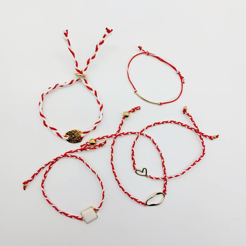 May include: Five red and white string bracelets with gold accents. The bracelets have different charms, including a heart, a circle, and a square.