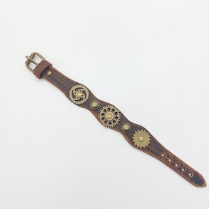 May include: A brown leather bracelet with a buckle closure and three brass gears. The gears are decorated with small brass studs.