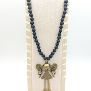 Può includere: Una collana di perline di lava nera con un ciondolo a forma di angelo dorato. Il ciondolo presenta una figura femminile stilizzata con ali e cuori sul suo vestito.