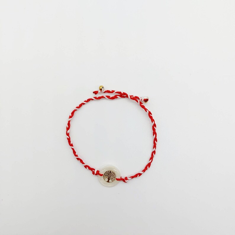 May include: Red and white braided string bracelet with a white round charm featuring a gold tree of life design.