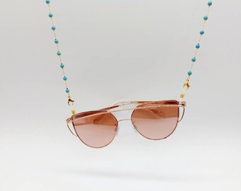 Könnte beinhalten: Eine goldene Kette mit türkisfarbenen Perlen, die eine Sonnenbrille in Roségold mit rosafarbenen Gläsern hält.
