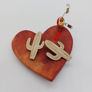 May include: A red heart-shaped wooden ornament with two wooden cactus cutouts. The ornament is decorated with a red ribbon and a small yellow and white bead.