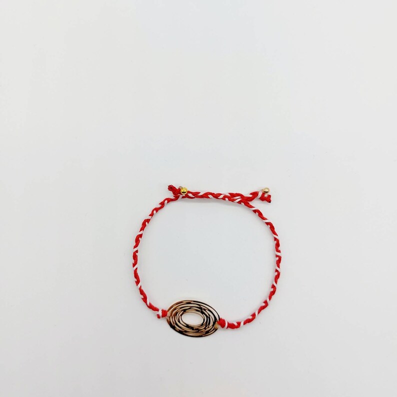 May include: A red and white braided string bracelet with a gold oval charm. The charm has a swirling design.