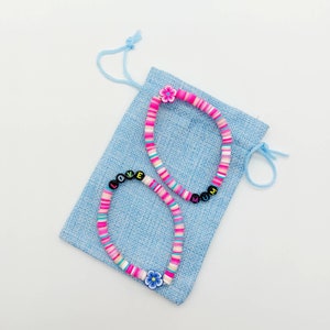 May include: Two colorful beaded bracelets with flower charms. One bracelet spells "LOVE" and the other spells "MUM". The bracelets are on a blue fabric pouch.