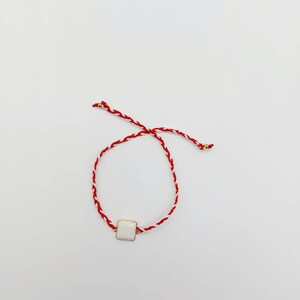 May include: A red and white braided string bracelet with a white square charm.