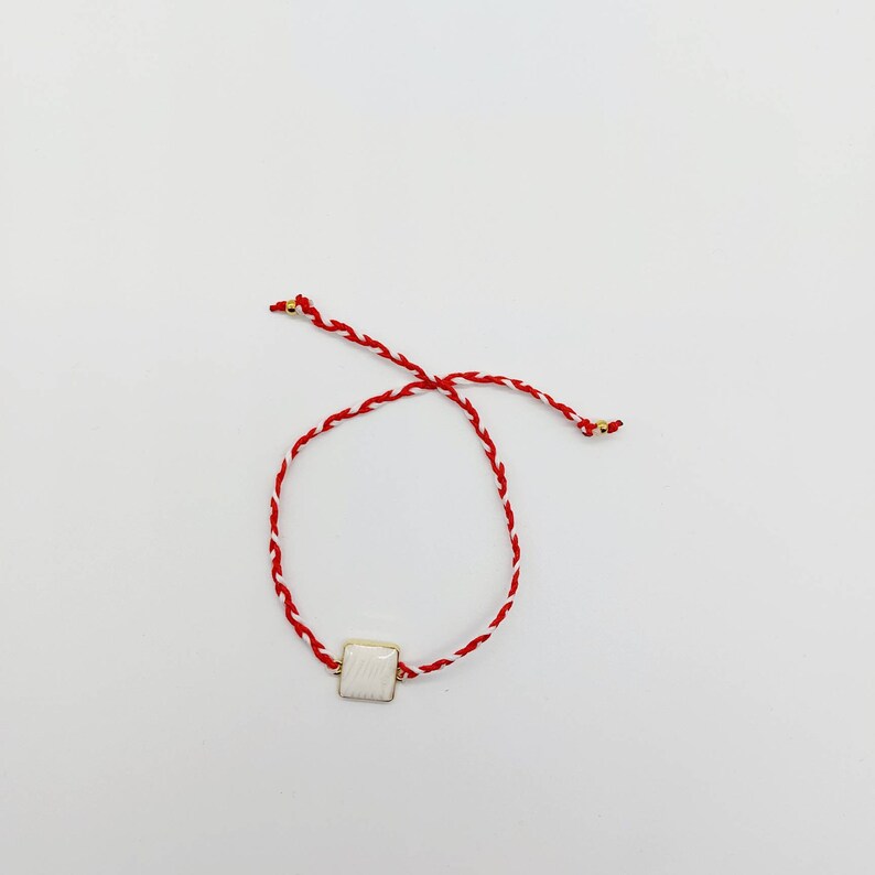 May include: A red and white braided string bracelet with a white square charm.