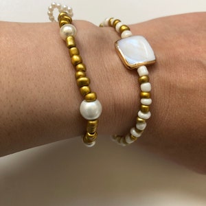 May include: Two bracelets, one with gold beads and white pearls, and the other with white beads and a white square charm with a gold rim.