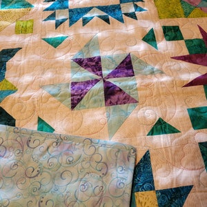 May include: A handmade quilt featuring a patchwork design with star and geometric patterns. The quilt showcases teal, purple, and green accents against a cream background. The backing has a blue and purple swirl pattern.
