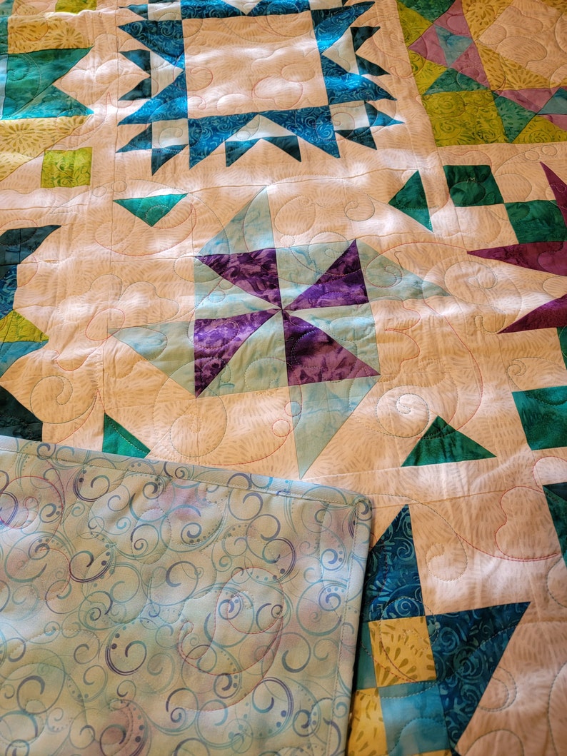 May include: A handmade quilt featuring a patchwork design with star and geometric patterns. The quilt showcases teal, purple, and green accents against a cream background. The backing has a blue and purple swirl pattern.