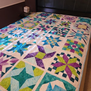 May include: A colorful quilt with a star pattern in shades of teal, purple, and green on a white background. The quilt is on a bed with a black headboard. The quilt is a patchwork design.