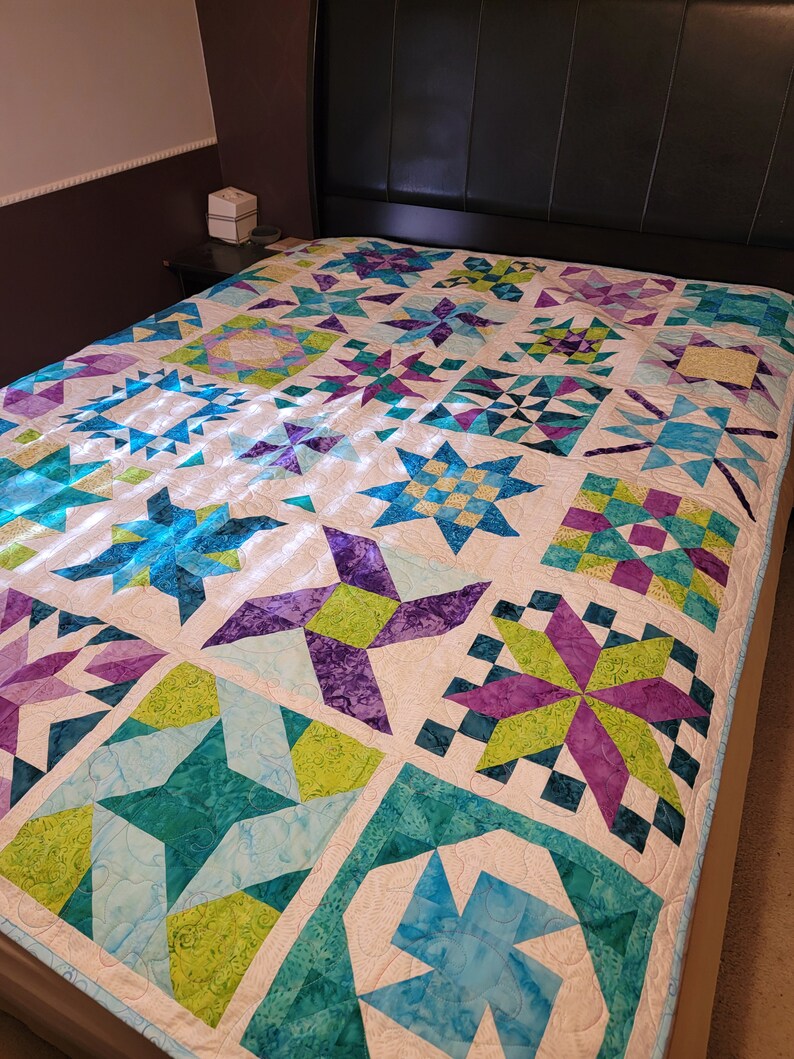 May include: A colorful quilt with a star pattern in shades of teal, purple, and green on a white background. The quilt is on a bed with a black headboard. The quilt is a patchwork design.