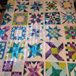 May include: A handmade quilt featuring a patchwork design with various star and geometric patterns. The quilt showcases a palette of teal, purple, and green hues against a cream background. The quilt is likely queen or king size.