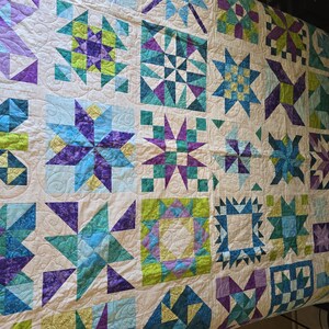 May include: A handmade quilt with a cream background and various geometric star patterns in shades of blue, purple, and green. The quilt is made of fabric squares and has a quilted texture.