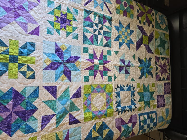 May include: A handmade quilt with a cream background and various geometric star patterns in shades of blue, purple, and green. The quilt is made of fabric squares and has a quilted texture.