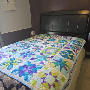 May include: A colorful quilt with a star pattern in shades of blue, purple, and green on a white background. The quilt is on a bed with a dark headboard. The quilt's border is light blue.