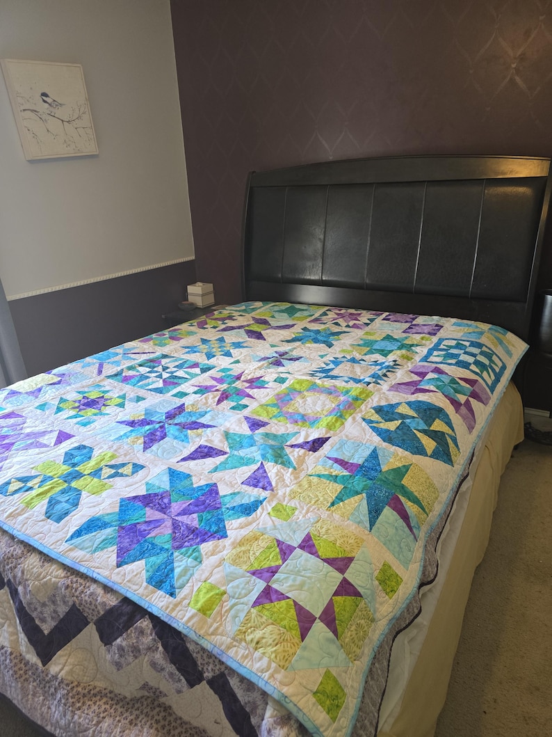 May include: A colorful quilt with a star pattern in shades of blue, purple, and green on a white background. The quilt is on a bed with a dark headboard. The quilt's border is light blue.