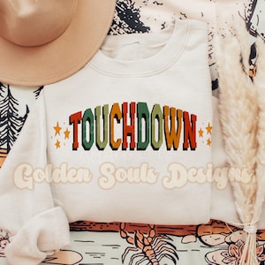 Puede incluir: Una sudadera blanca con el texto "TOUCHDOWN" en letras retro de colores. Las letras están delineadas en negro y tienen un aspecto vintage. Hay dos pequeñas estrellas a cada lado del texto.