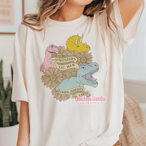 Könnte beinhalten: Ein weißes T-Shirt mit einem grafischen Design mit Dinosauriern, Blumen und dem Text "Dinosaurs Eat Man, Woman Inherits The Earth, Golden Souls Design Studio".