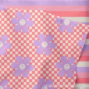 May include: A pink and white checkered fabric with a repeating pattern of purple flowers with smiley faces. The fabric is folded over a pink and white striped fabric.