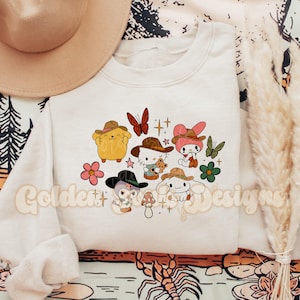 Könnte beinhalten: Cremefarbenes Sweatshirt mit einem farbenfrohen Aufdruck von Cartoon-Charakteren mit Cowboyhüten, umgeben von Blumen und Schmetterlingen. Der Text "Golden Designs" ist unten aufgedruckt. Ein beiger Hut ist oben links zu sehen.