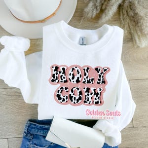 Könnte beinhalten: Weißer Pullover mit einem pinkfarbenen und schwarzen Kuhmuster-Design, auf dem in einer Retro-Schriftart "HOLY COW" steht. Der Pullover liegt auf einer Holzoberfläche mit einem weißen Hut und einer blauen Jeans.