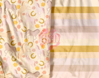 May include: A white fabric with a repeating pattern of horses, horseshoes, and flowers in shades of pink, yellow, and brown. The fabric is next to a white fabric with horizontal stripes in shades of beige and gold.