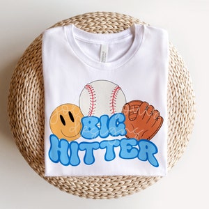 Könnte beinhalten: Weißes T-Shirt mit einem blauen, gelben und orangefarbenen Grafikdesign mit einem Baseball, einem Handschuh und einem Smiley. Der Text "BIG HITTER" ist in blauen Buchstaben geschrieben.