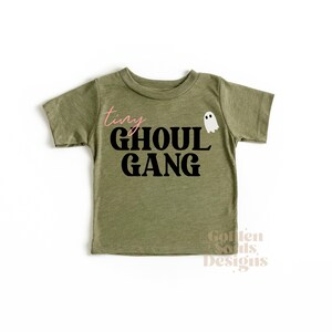 Può includere: Una maglietta verde a maniche corte con il testo "tiny ghoul gang" in lettere nere e un'immagine di un fantasma bianco.