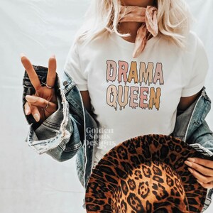 K&ouml;nnte beinhalten: Wei&szlig;es T-Shirt mit dem Text "DRAMA QUEEN" in tropfenden pinken, orangen und gelben Buchstaben.