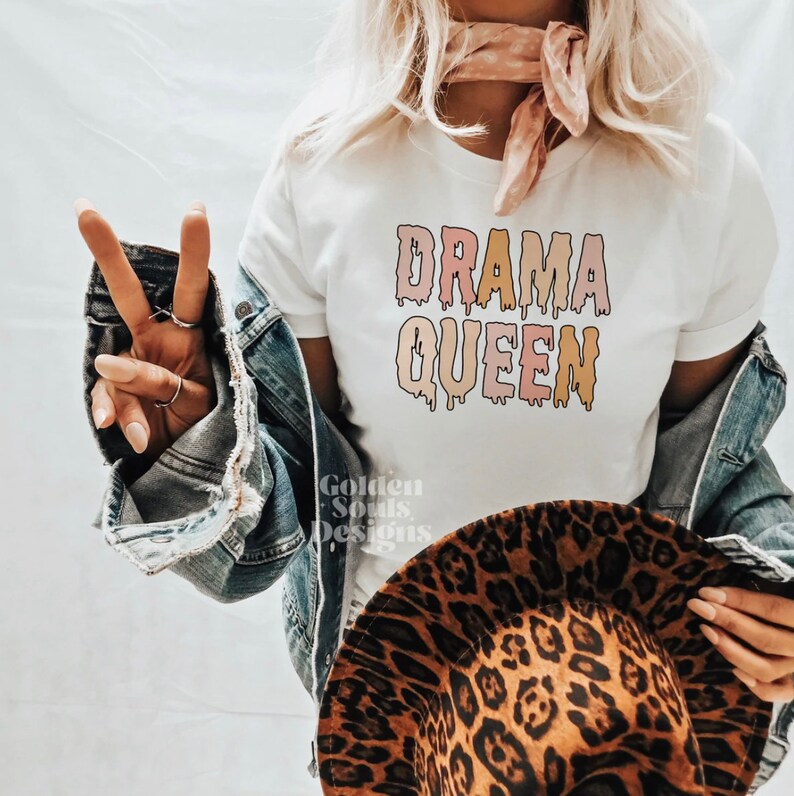 K&ouml;nnte beinhalten: Wei&szlig;es T-Shirt mit dem Text "DRAMA QUEEN" in tropfenden pinken, orangen und gelben Buchstaben.
