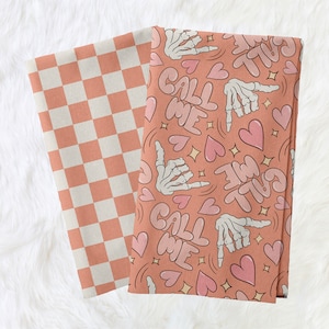 May include: Two fabric squares, one with a checkered pattern in peach and white, and the other with a pink background and a repeating pattern of a skeleton hand making a call me sign, hearts, and the words "Call Me Twice" in a bubble font.