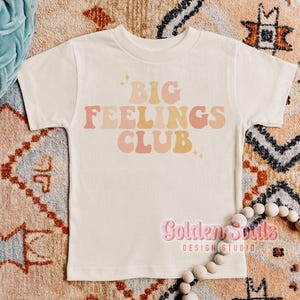 Könnte beinhalten: Cremefarbenes T-Shirt mit dem Text "BIG FEELINGS CLUB" in Retro-Schrift. Der Text ist in Rosa-, Pfirsich- und Gelbtönen gehalten. Das Shirt wird auf einem gemusterten Teppich präsentiert.