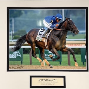 May include: A framed photograph of a racehorse and jockey in action. The horse is dark brown with a white marking on its face, and the jockey wears blue and white. The image includes text that reads "SOVEREIGNTY" and "SARATOGA".