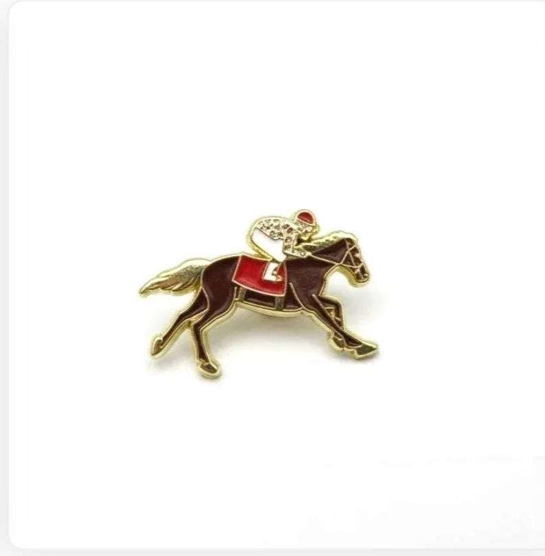 May include: A gold-coloured pin depicting a horse and jockey in a racing position. The horse is brown with a red saddle, and the jockey wears a red cap and a white and gold patterned uniform. This pin is likely a collectible or equestrian-themed jewellery.