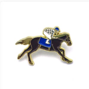 May include: A gold-coloured pin depicting a racehorse and jockey. The horse is dark brown with a blue saddle, and the jockey wears a white and checkered uniform with a blue helmet. The pin has a detailed design and is set against a white background.