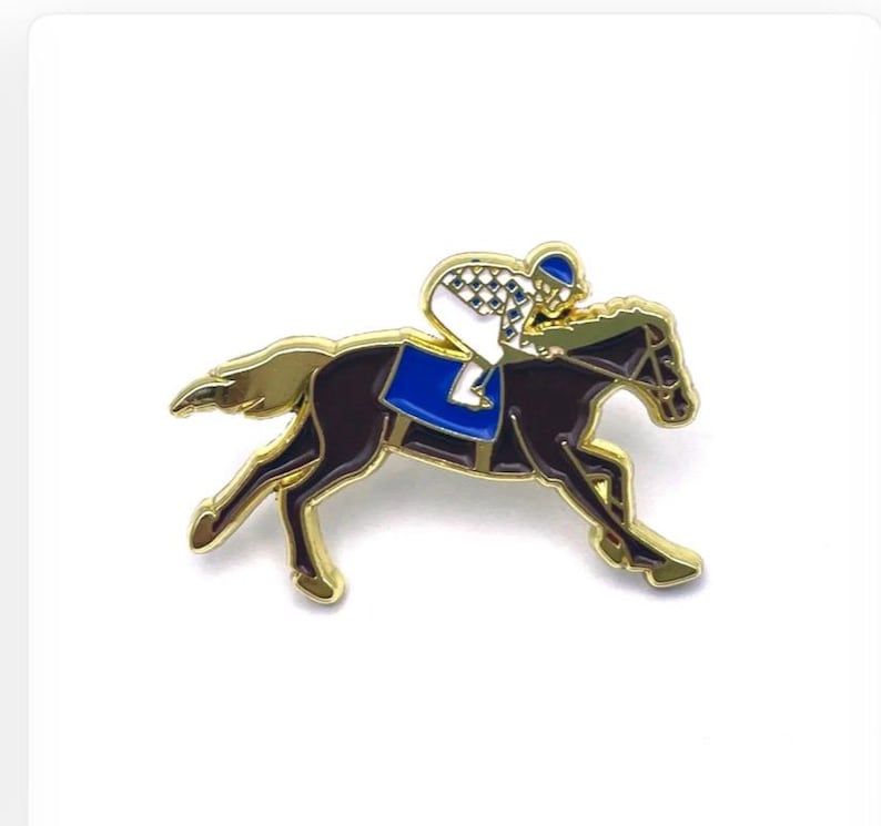 May include: A gold-coloured pin depicting a racehorse and jockey. The horse is dark brown with a blue saddle, and the jockey wears a white and checkered uniform with a blue helmet. The pin has a detailed design and is set against a white background.