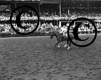 May include: A black and white photo of a horse and jockey racing on a dirt track. The horse is running towards the right side of the image. The jockey is wearing a white uniform and a helmet. There are people in the background watching the race.