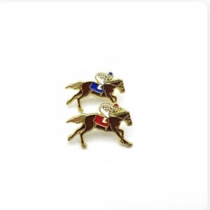May include: Two gold-coloured horse racing pins. Each pin depicts a brown horse with a jockey wearing a helmet and silks. One jockey has a blue saddle blanket, the other a red one. The pins are set against a white background.