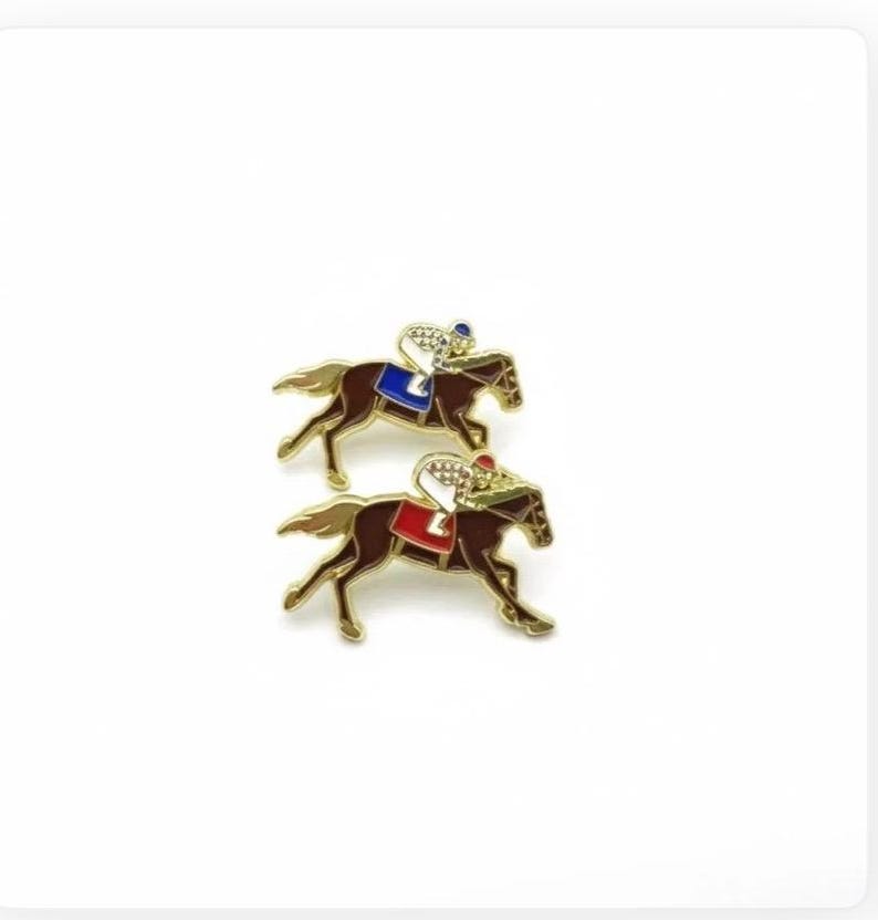 May include: Two gold-coloured horse racing pins. Each pin depicts a brown horse with a jockey wearing a helmet and silks. One jockey has a blue saddle blanket, the other a red one. The pins are set against a white background.