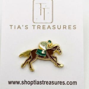 May include: A gold-coloured pin depicting a brown horse with a jockey in green and white. The pin is on a white card with the text "TIA'S TREASURES" and "www.shoptiastreasures.com".