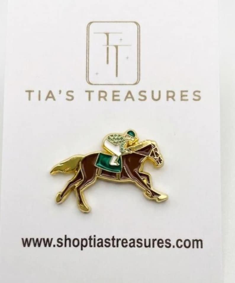 May include: A gold-coloured pin depicting a brown horse with a jockey in green and white. The pin is on a white card with the text "TIA'S TREASURES" and "www.shoptiastreasures.com".