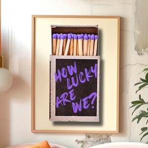 May include: A framed print of a matchbox with the text "HOW LUCKY ARE WE?" written in purple on the side. The matchbox is filled with matches with purple heads.