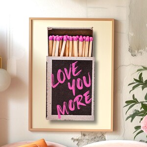 May include: A framed print of a matchbox with pink matches and the text "LOVE YOU MORE" written in pink on a black background.