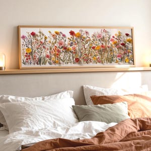 May include: A rectangular framed floral print showcasing a colourful arrangement of wildflowers in yellow, red, pink, and purple hues. The artwork is positioned above a bed with white and orange bedding. Two small lamps with warm light are on either side.